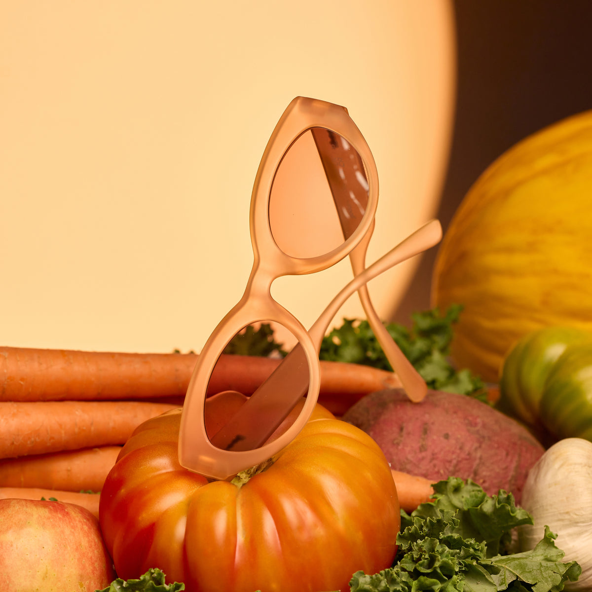 A pair of folded translucent peach cat-eye sunglasses sitting on top of harvested vegetables against a moon backdrop.