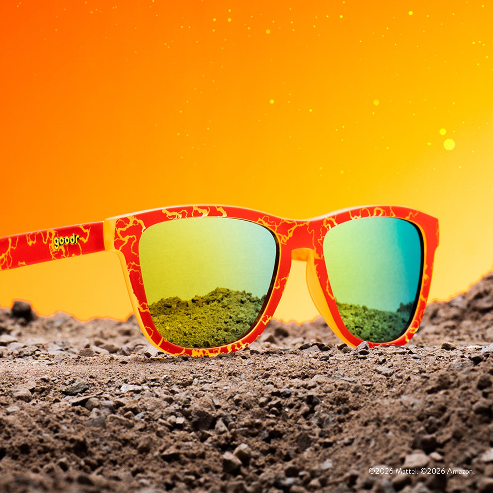 Three-quarter angle view of red and yellow lightning bolt-patterned sunglasses with reflective gold lenses, set against an orange & gravel backdrop.