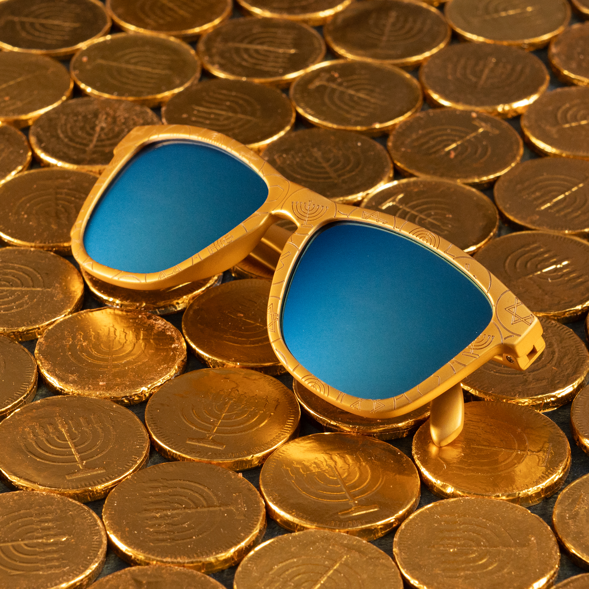 Folded golden sunglasses featuring a gelt print and blue reflective lens, placed on top of wrapped chocolate gelt coins.