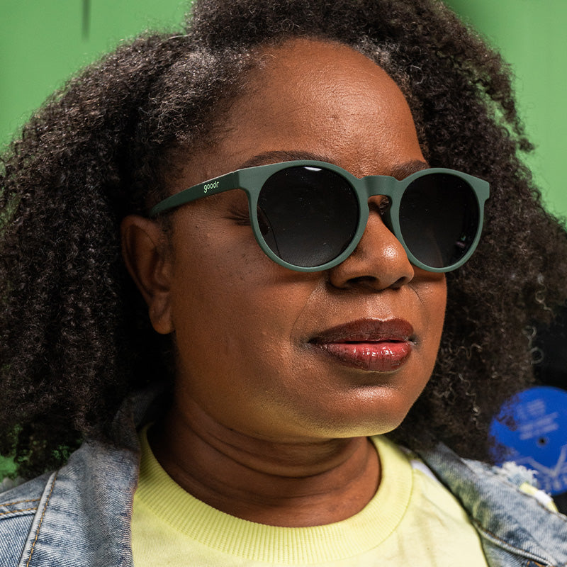 A woman in dark green round sunglasses with non-reflective gradient green lenses looks off to the side confidently.