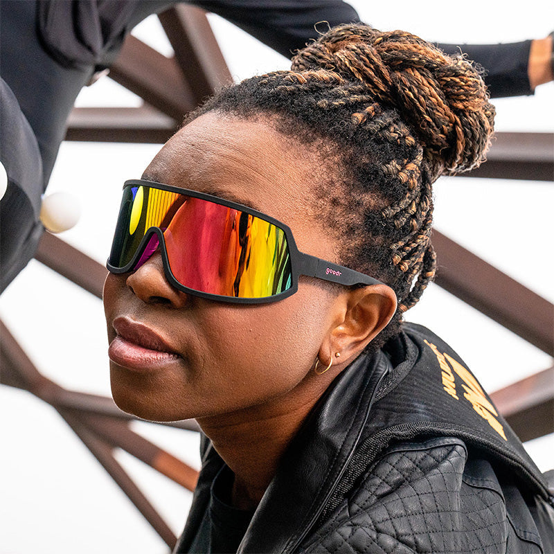 A stunt woman wearing black wraparound sunglasses with a pink reflective lens looks into the distance, a bridge behind her.
