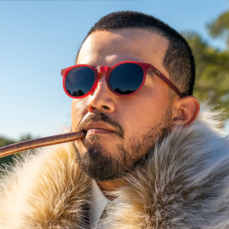 A man wearing round burgundy sunglasses with brown lenses and a large furry collar smokes a pipe on a golf course.