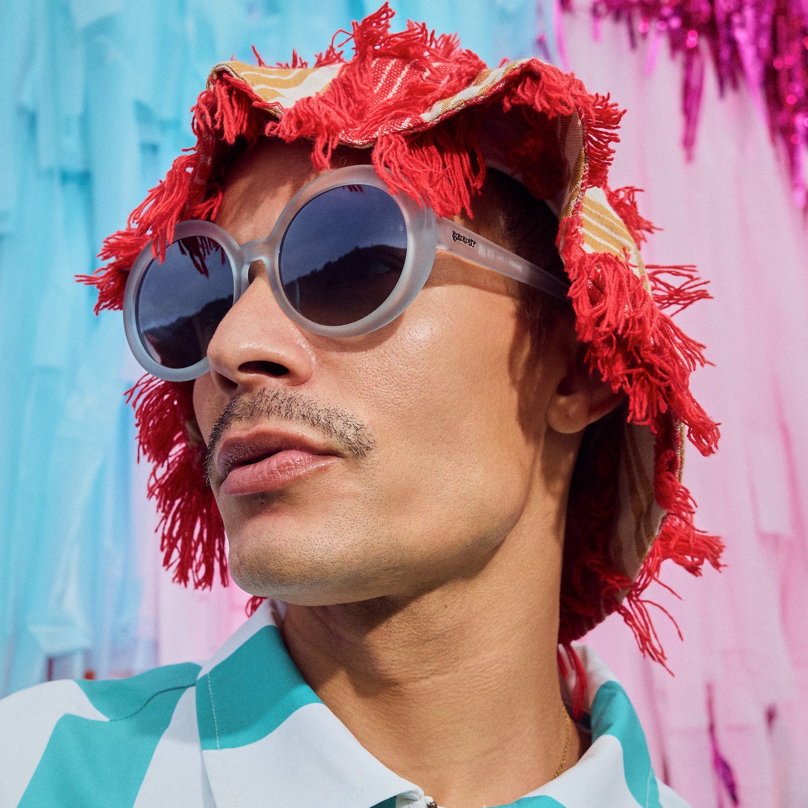Side profile of a man wearing a red hat and oversized round sunglasses with translucent blue frames and gradient lenses against a pastel backdrop.