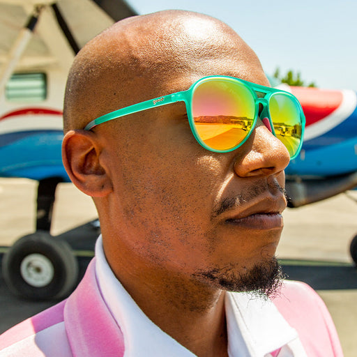 A man in teal aviator sunglasses with pink reflective lenses looks out into the distance, a small airplane behind him.