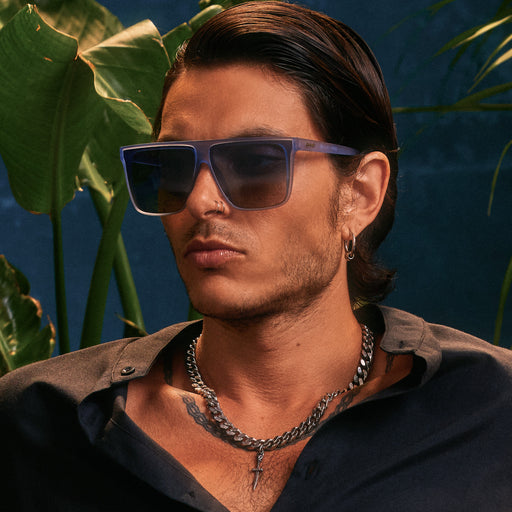 A handsome young man with dark hair poses in oversized, dusty blue Fly G shades against a deep blue backdrop and palm fronds.