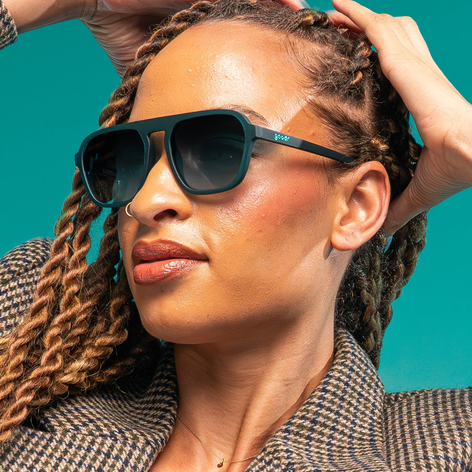 A woman with copper twist braids in a houndstooth coat is casually pulling her hair back, revealing a pair of translucent teal Retro Gs.