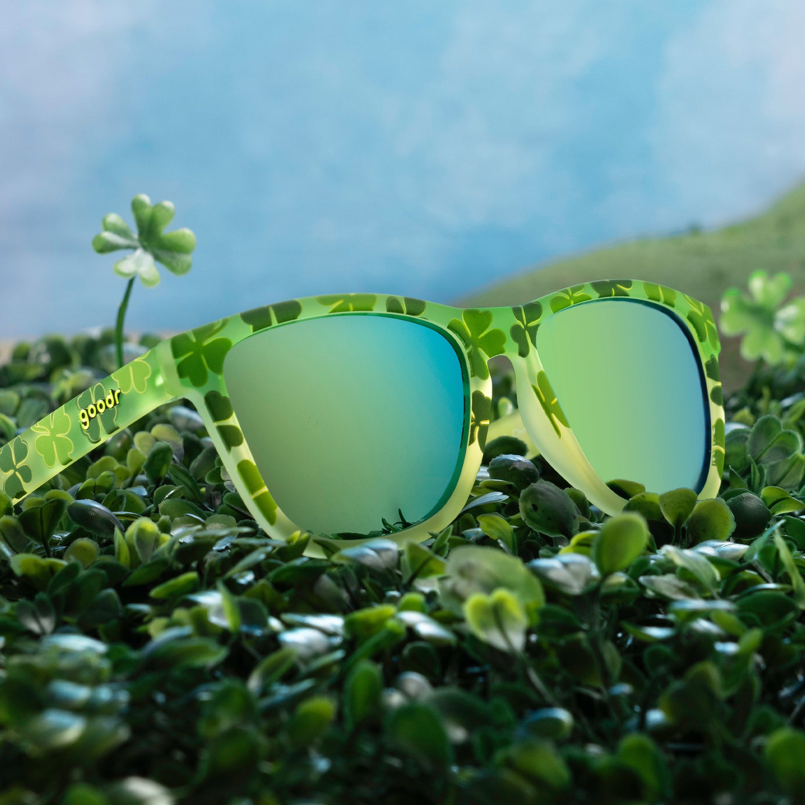 Three-quarter angle view of green shamrock print sunglasses w/ green+gold reflective lenses in a field of shamrocks.