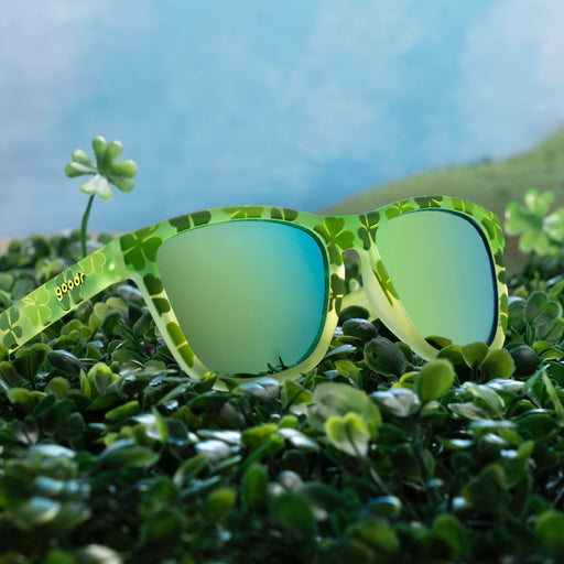 Three-quarter angle view of green shamrock print sunglasses w/ green+gold reflective lenses in a field of shamrocks.