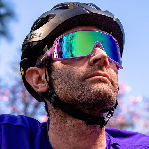 A cyclist wearing a bike helmet and wraparound purple sunglasses with a reflective green lens looks ahead on a sunny day.