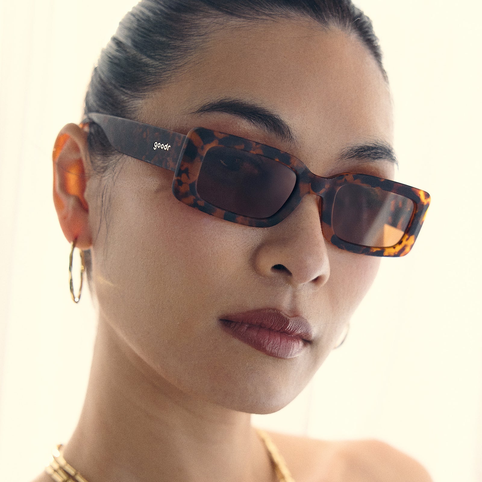 A woman with a sleek bun wears a pair of tortoiseshell rectangular sunglasses with brown polarized lenses.