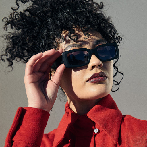 A woman poses, holding and wearing black rectangular sunglasses with black polarized lenses.