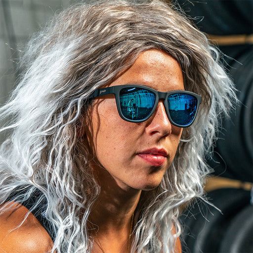 A woman in a curly gray mullet wig looks off to the side wearing black sunglasses with blue reflective lenses.