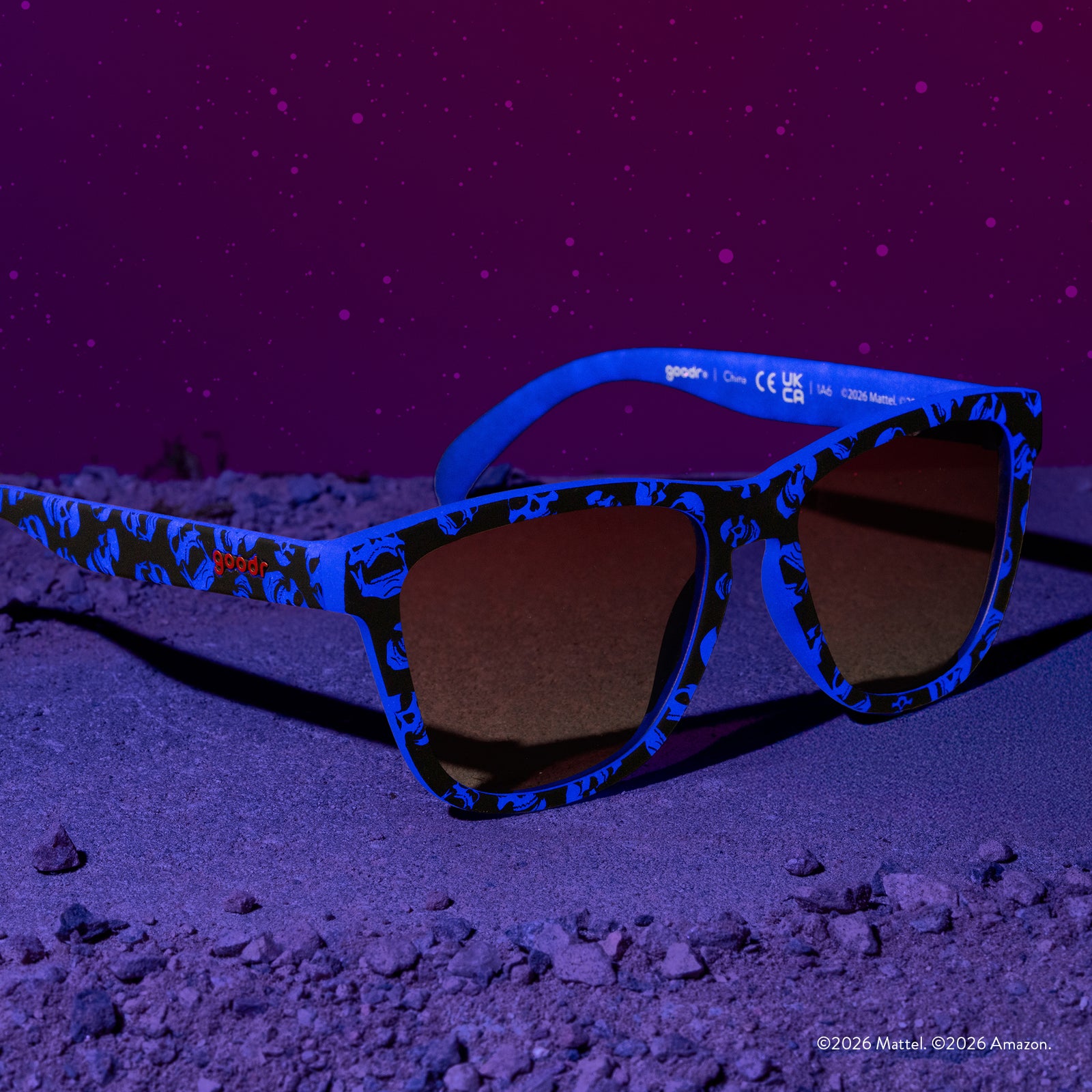 Three-quarter angle view of blue and black sunglasses featuring a Skeletor pattern and rose gradient lenses, set against a low-lit purple background.