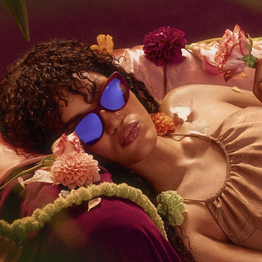 A woman lounges, posing in translucent purple cat-eye sunglasses with purple reflective lenses.