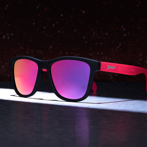 Three-quarter angle view of black and pink sunglasses illuminated by a spotlight against a dark, starry background.