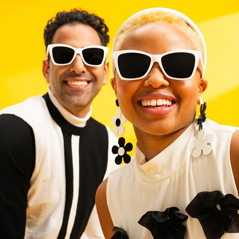 A man and woman in retro black and white outfits smile in white angled cat-eye sunglasses with black lenses.