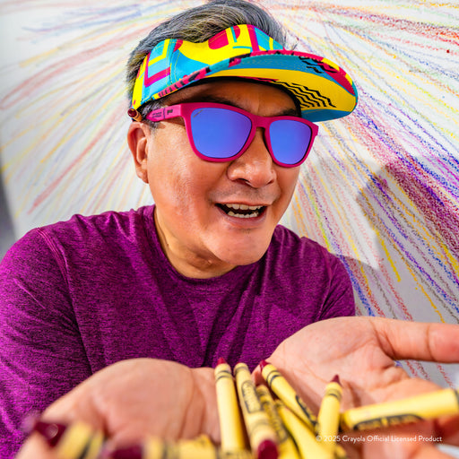 A silver-haired man in a colorful visor wearing Mulberry-colored Crayola sunglasses holds a handful of Mulberry crayons