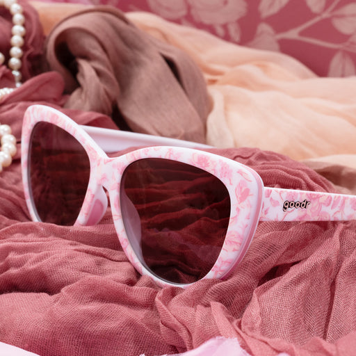 Three-quarter angle view of pink and white sunglasses with a floral toile pattern and rose gradient lenses on top of different pink-colored fabric.