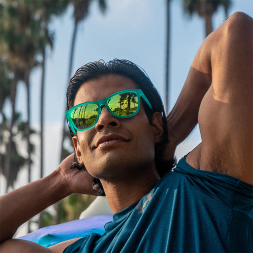 A smirking muscular man wears teal sunglasses with teal reflective lenses and flexes his biceps, palm trees in the distance.