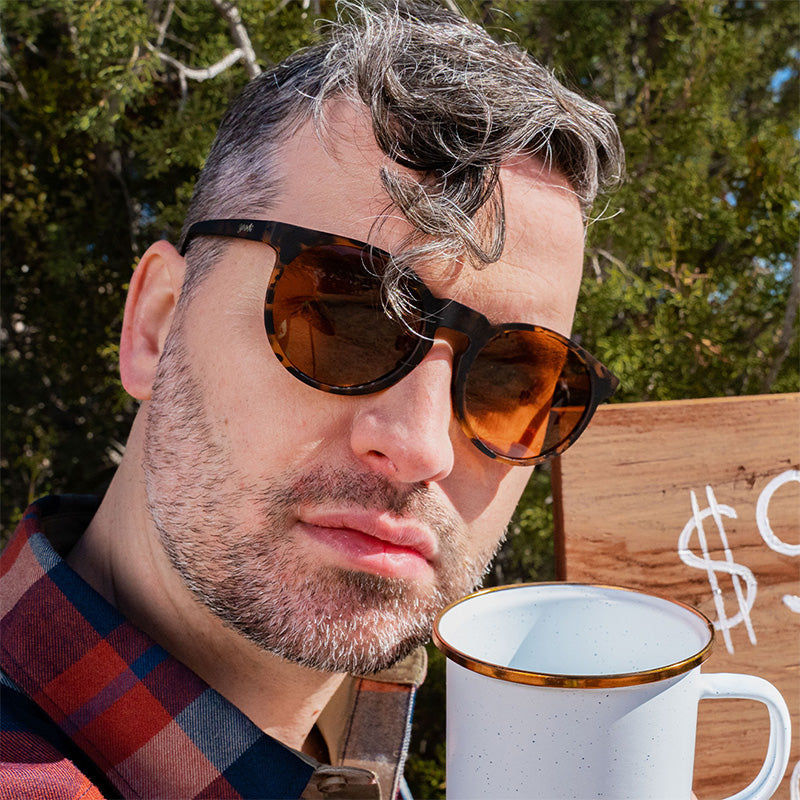 A hipster barista judgmentally glares ahead wearing brown tortoiseshell sunglasses with brown non-reflective lenses.