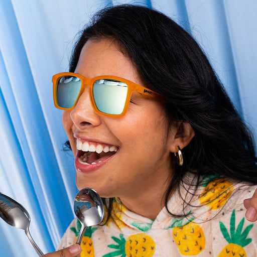 A woman wearing small square-shaped orange sunglasses with mirrored reflective lenses smiles gleefully while holding two spoons.