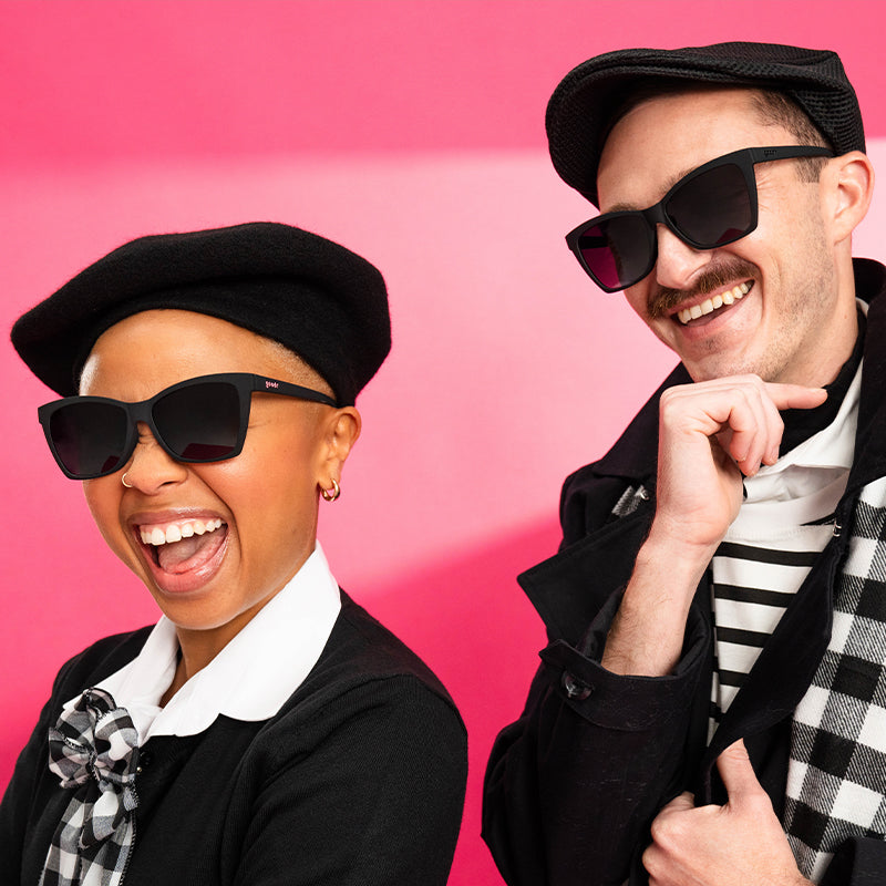 A man and woman in stylish black and white outfits with black angled cat-eye sunglasses with black lenses laugh.
