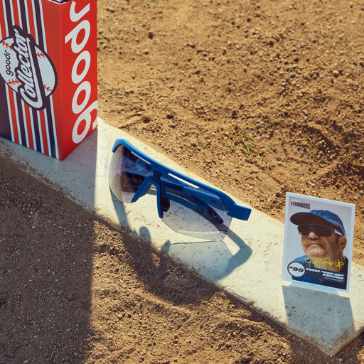 Front view of blue sunglasses w/ blue gradient lenses, custom packaging & trading card lined up on a dirt ballfield.
