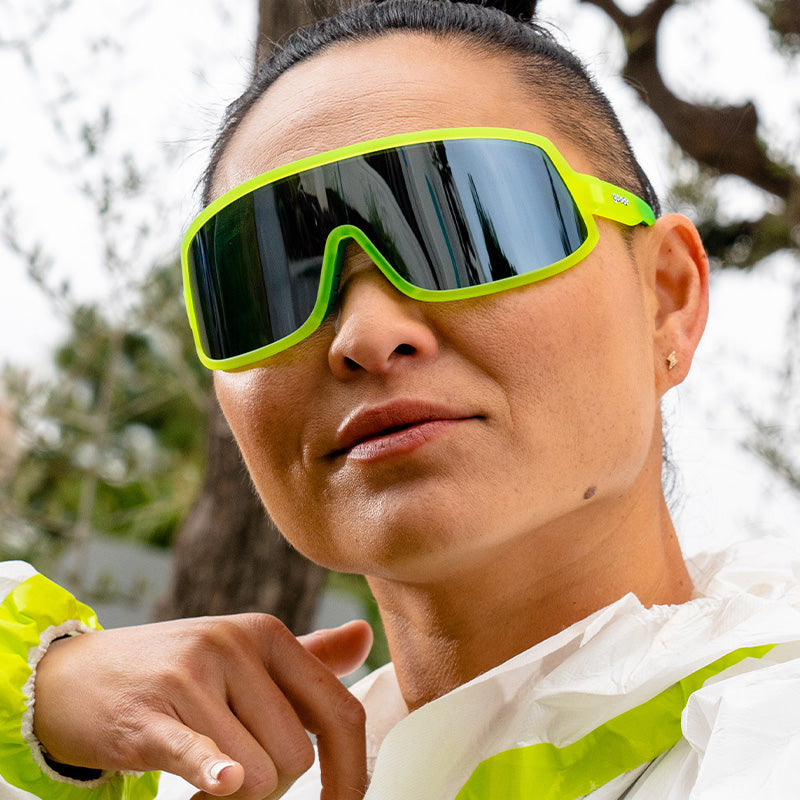 A woman wearing a hazmat suit and neon yellow sunglasses with a reflective gray lens looks fiercely ahead.