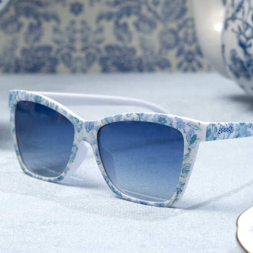 Three-quarter angle view of blue and white angular cat-eye sunglasses with a floral toile and blue gradient lenses on top of a blue velvet tablecloth.