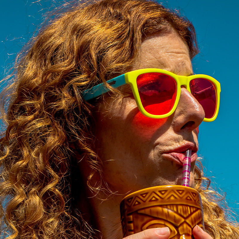 A woman wearing light blue and yellow sunglasses with square rose lenses sips a tropical drink on a sunny day.