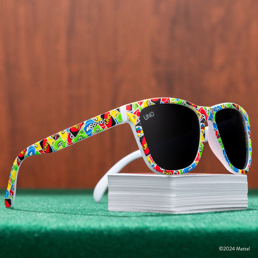 Three-quarter view of a pair of UNO shades perched atop a deck of UNO cards, on a green game table in front of wood paneling.