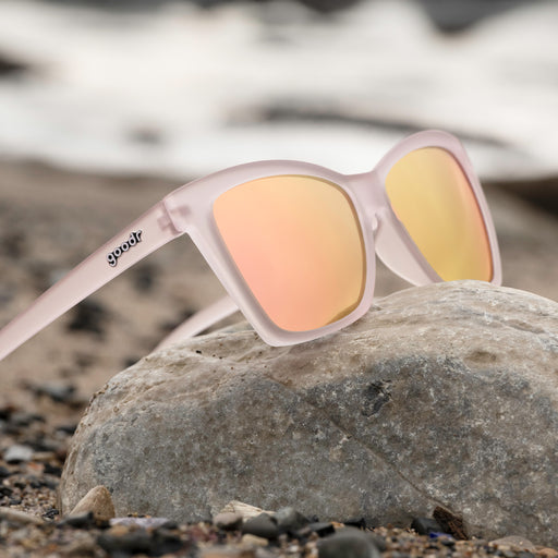 Three-quarter view of translucent pink cat-eye glasses with light pink reflective lenses propped on a gray rock, seaside.