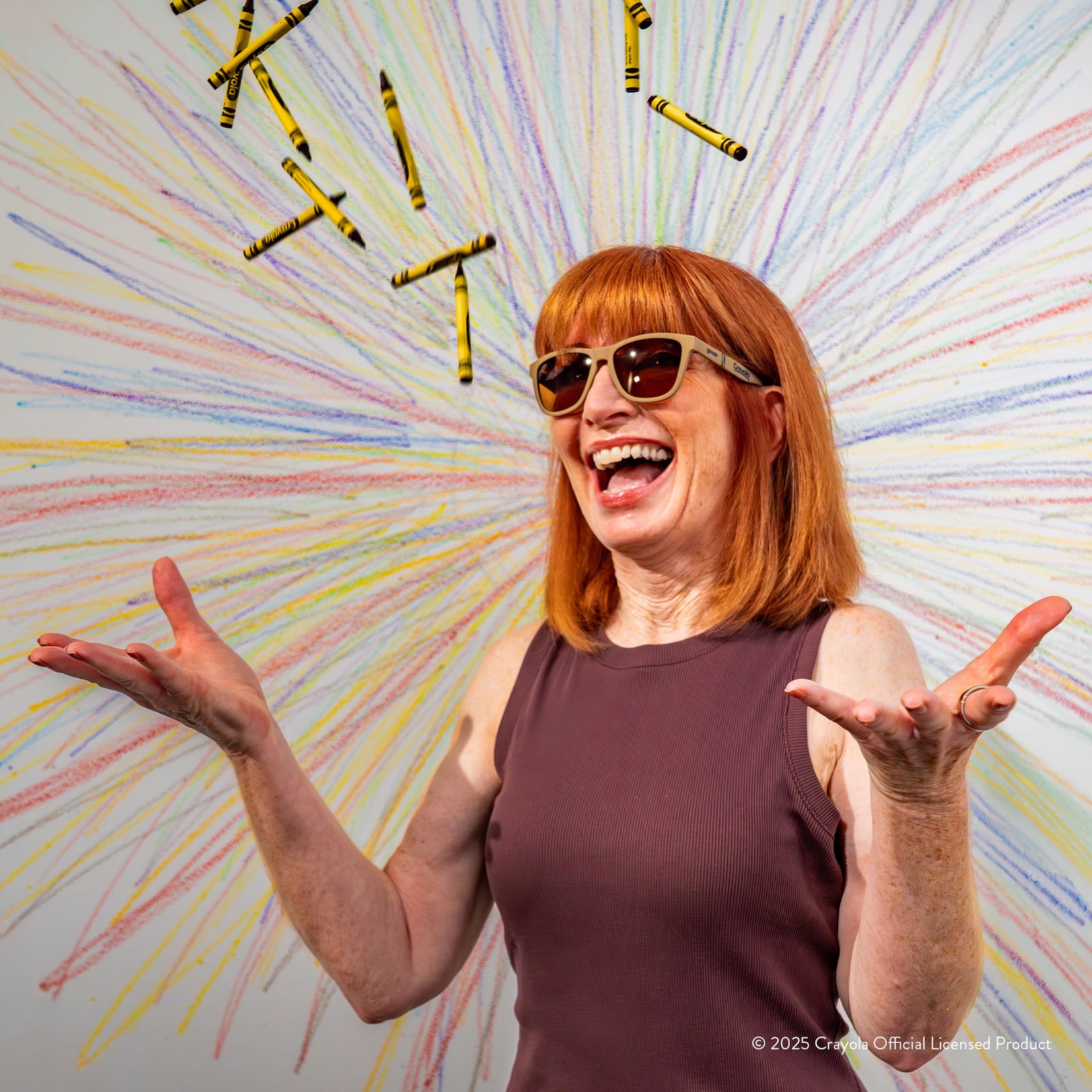 A laughing redheaded woman wearing Raw Umber-colored Crayola sunglasses joyfully tosses Raw Umber crayons into the air