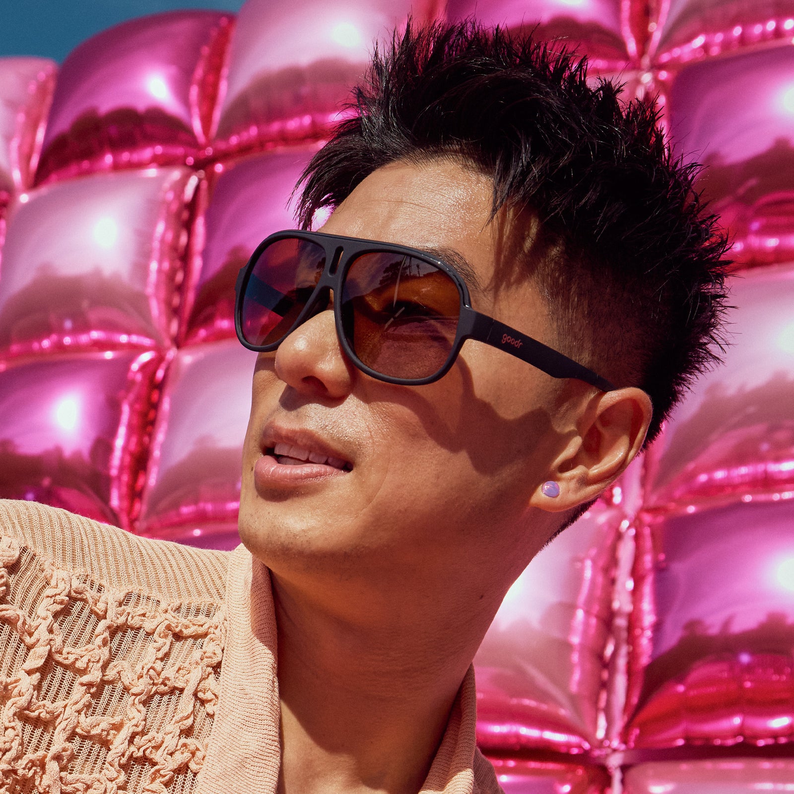 Side profile of a man wearing black aviator sunglasses with amber gradient lenses in front of a giant shiny pink balloon wall.
