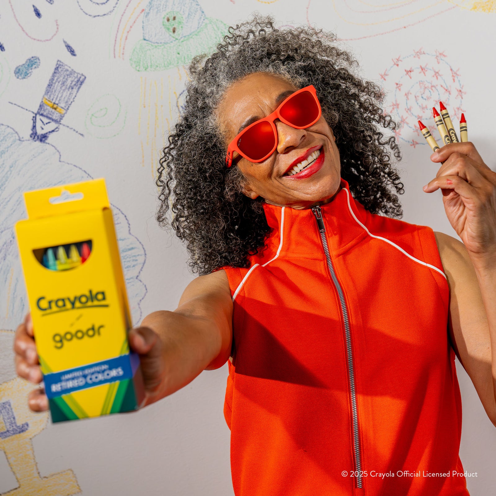 A smiling older woman wearing Orange Red Crayola shades & a matching top holds up crayons and the Crayola box packaging