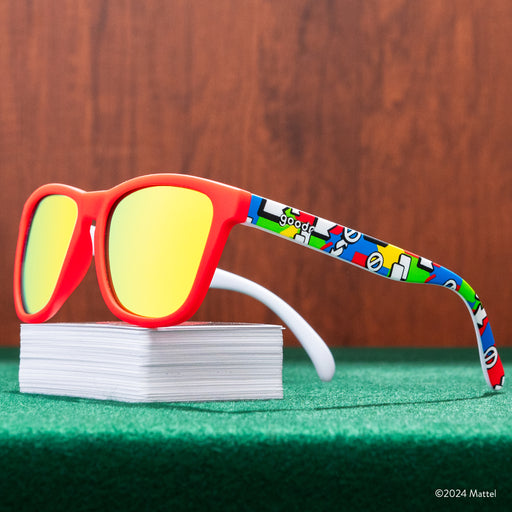 Three-quarter view of a pair of UNO shades perched atop a deck of UNO cards, on a green game table in front of wood paneling.