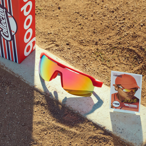 Front view of red sunglasses w/ burnt orange reflective lenses, custom packaging & trading card on a pitcher's mound.