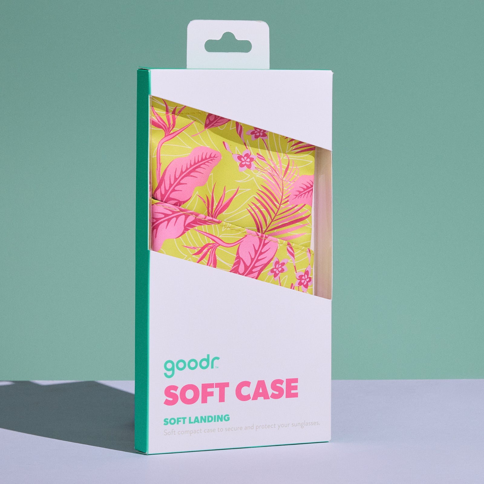 A white product box containing the pink and yellow Soft Landing sunglasses case is displayed on a white tabletop with a sage green backdrop.