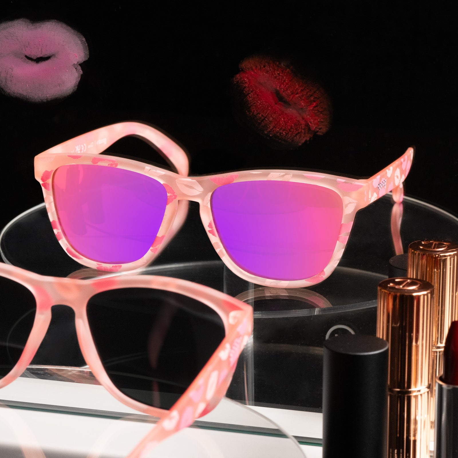 Three-quarter angle view of pale pink sunnies featuring rose reflective lenses and a flirty all-over-kiss print sitting on a black vanity top.
