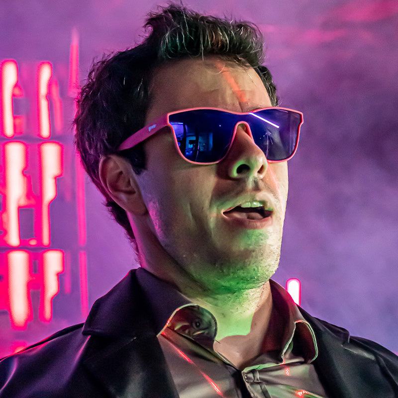 A man in hot pink futuristic sunglasses with a single reflective purple lens intensely stares off in a foggy light-up bar.