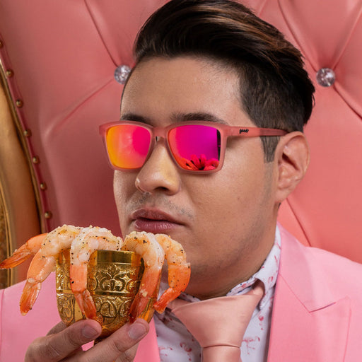 A man wearing small square-shaped pink sunglasses sits on his throne while drinking from a golden goblet filled with shrimp.
