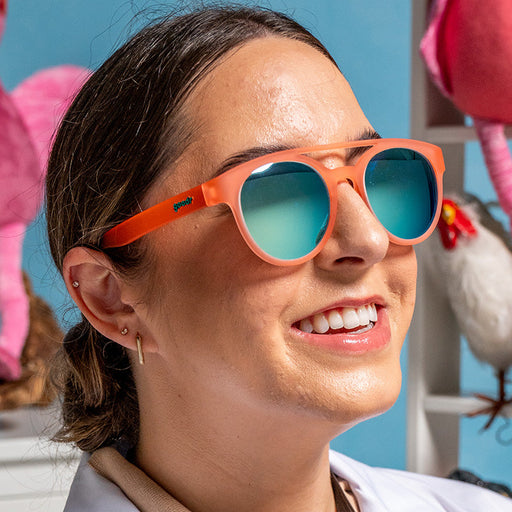 A woman wearing round orange sunglasses with a double bridge and teal reflective mirrored lenses smiles with glee in her room full of stuffed animals.