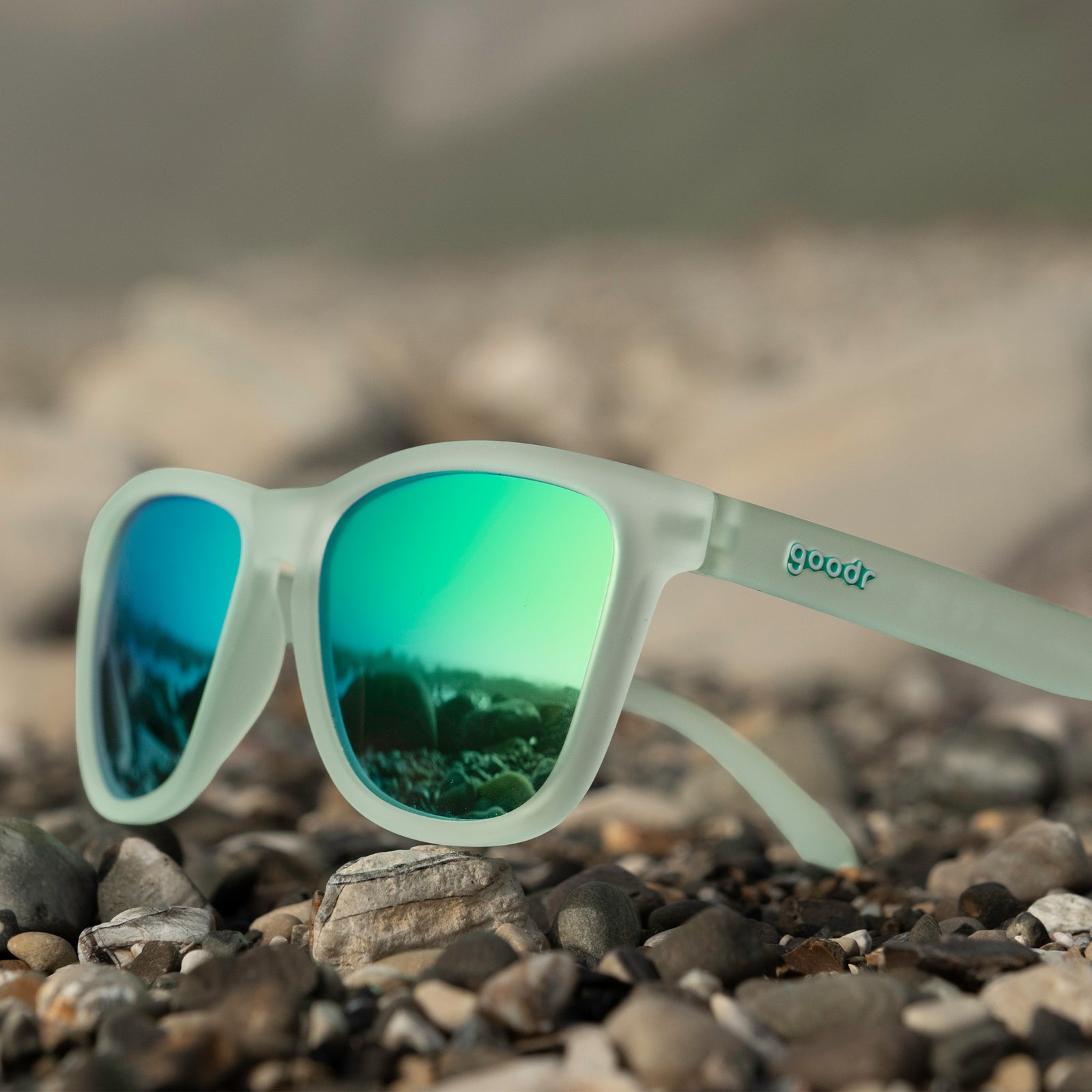 Three-quarter view of translucent green OG sunglasses with green-blue reflective lenses lying atop beach pebbles.