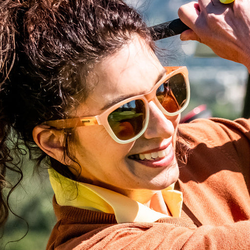 A woman wearing brown-to-white gradient sunglasses swings a golf club, smiling in a ray of sunshine.