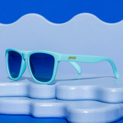 Three-quarter angle view of teal sunglasses with blue-green gradient polarized lenses resting on a cool blue wavy display representing antacid relief.