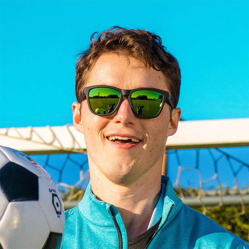 A laughing man wearing black sunglasses with green reflective lenses looks ahead, holding a soccer ball in front of a goal.