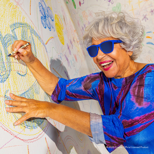 A happy older woman with gray hair wearing Violet Blue Crayola sunglasses colors on a white wall with colorful drawings