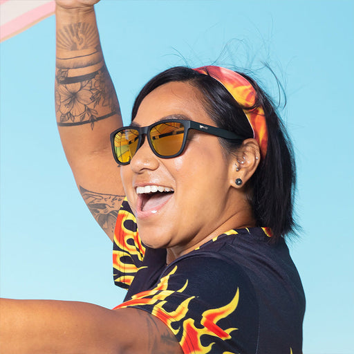 A woman in black sunglasses with amber reflective lenses wearing a flame-patterned shirt and headband laughs.