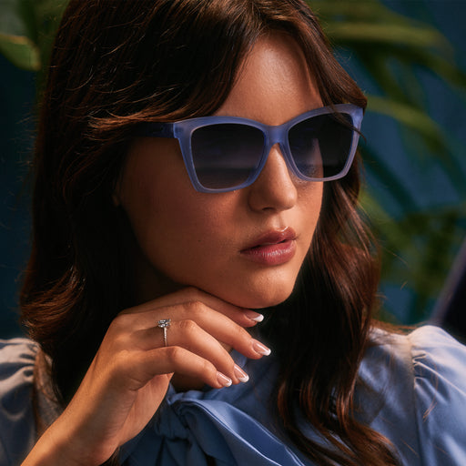 A woman in translucent blue Pop Gs rests her chin thoughtfully on her hand, blue backdrop and palm fronds in the background.