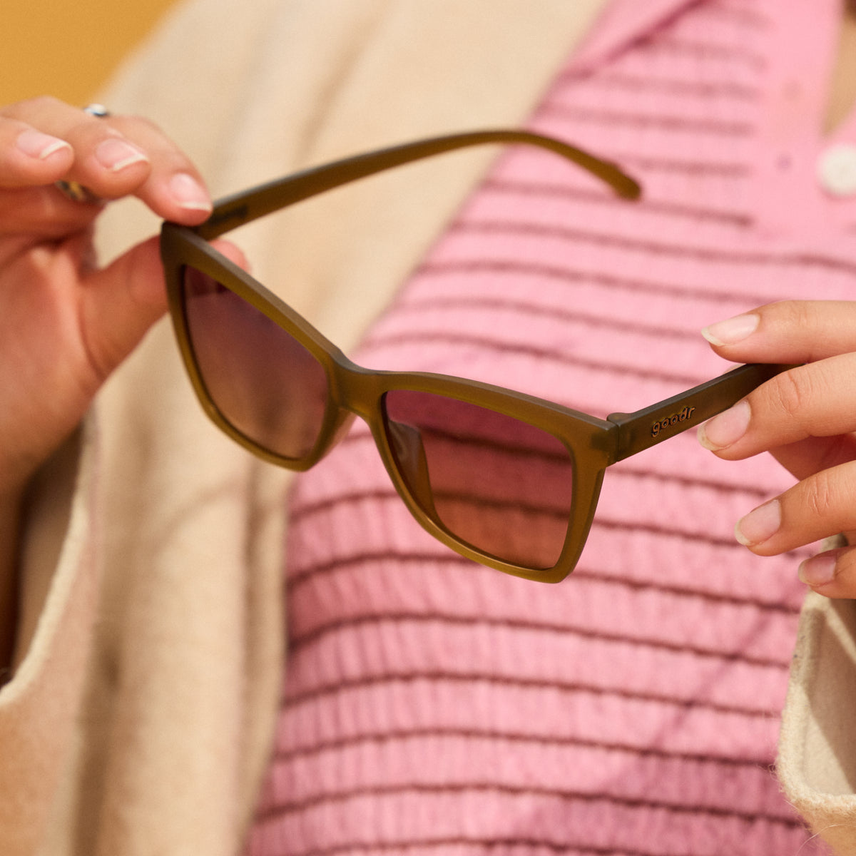Close-up of a woman’s hands holding a pair of translucent ochre sunglasses in front of her pink and cream outfit.